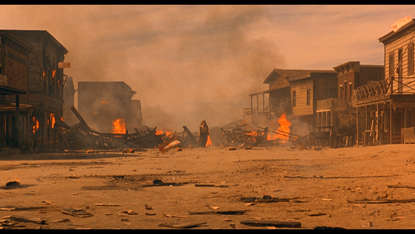A lone figure stands among burning buildings in a dusty Old West town.